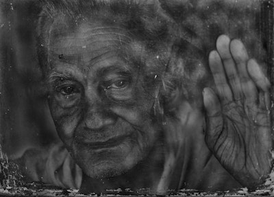 Elderly person looking out through a weathered window, hand raised against the glass.